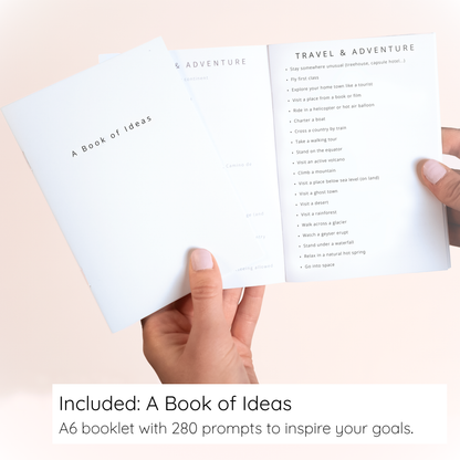 Hand holding an open booklet titled 'A Book of Ideas' with prompts on a light pink background.
